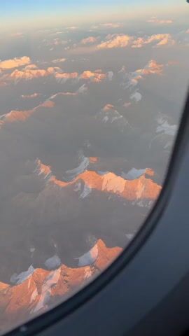 An aerial view from an airplane window shows snow-capped mountain ranges illuminated by the warm glow of sunrise or sunset. The mountains are rugged and jagged, with sharp peaks and deep valleys. Patches of snow and ice gleam on the upper slopes and summits, contrasting with the darker, rocky terrain below. The sky above is a clear blue, transitioning to a hazy orange and pink band near the horizon where the sun is setting or rising. Wispy clouds are scattered across the sky, catching the light and adding a soft, ethereal quality to the scene. The overall impression is one of vastness, natural beauty, and the serene solitude of high altitudes.