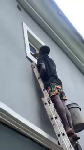 A person is on a ladder painting a window frame. The person is wearing a black shirt and colorful shorts and standing on an aluminum ladder.  A bucket is attached to one of the lower rungs of the ladder. The building being painted is light gray with white trim. The window is triangular. The video is shot from a low angle.