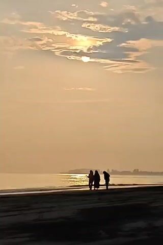 The video shows a landscape of a beach, with three people walking along the water's edge. The people are silhouetted against the sunrise or sunset, with the sun partially obscured by clouds. The sky is a light orange color, and the water reflects the sunlight. The sand on the beach is a darker color.