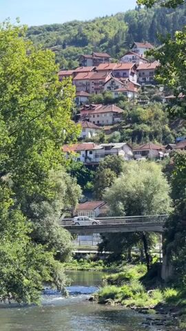 A small, tree-lined river flows in the foreground, with a concrete bridge spanning across it. On the bridge, a white car drives from left to right. Beyond the bridge, a hillside is dotted with numerous houses, each with red-tiled roofs. The houses are nestled among green trees and appear to be part of a small village or town. The sky above is a clear blue. The scene is tranquil and rural, with the natural landscape and the human settlement integrated into the hillside.