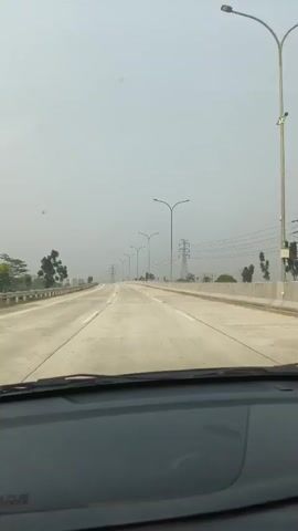 The video shows a first-person view from inside a vehicle traveling on a highway. The highway is wide, with multiple lanes, and appears to be made of concrete. There are streetlights lining the side of the road, and in the distance, there are electrical pylons. The sky is overcast and hazy, and the overall visibility is reduced. The dashboard of the vehicle is visible in the foreground, and the windshield offers a clear view of the road ahead. There are no other vehicles or people visible in the immediate vicinity. The environment appears to be a developed area, likely an urban or suburban outskirts, given the infrastructure present.