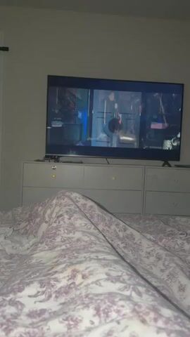 The video shows a bedroom scene. In the foreground, a person is lying in bed covered by a patterned duvet. A television is mounted on a dresser in the background, displaying a scene with two people. The camera then pans to the right, revealing a man sitting on the bed, holding a black object with a yellow tip. He is wearing a blue t-shirt and appears to be looking at the camera. Behind him, white curtains are drawn over a window. A small, colorful object sits on a table to his right. The camera then pans back to the left, showing a dresser with drawers and a sliding door in the background. The video has a dark and somewhat blurry quality.