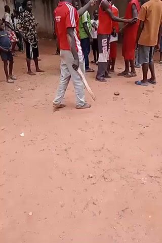A group of young men, mostly wearing red shirts and shorts, are gathered on a dirt-covered area. One individual, wearing a red basketball jersey with the number 13 and the name "HARDEN" on the back, is interacting with another person. There is a wooden stick lying on the ground near a small object, possibly a ball. A wall with barbed wire is visible in the background. The atmosphere appears to be informal, possibly a casual game or gathering.