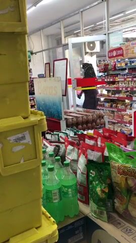 The video captures a retail environment, likely a convenience store or small supermarket, viewed from a low angle. In the foreground, large, bright yellow plastic storage containers are stacked high on the left side of the frame, partially obscuring the view of the interior. To the right of the containers, several bottles of green liquid, appearing to be a carbonated beverage, are arranged on a shelf. Behind these bottles, various bagged food products, including snacks and what appear to be cereal boxes, are displayed. A woman, dressed in dark clothing and a headscarf, is visible further back in the store, pushing a red shopping cart containing a yellow box. She appears to be walking towards the right of the frame. The store interior features aisles of shelves stocked with products, overhead lighting, and a ceiling with visible infrastructure. Signage is present, including one that reads "Beras," which translates to "rice" in Indonesian, and another red and white sign that says "Bread Corner." The overall impression is a busy retail setting with merchandise on display and a customer making a purchase or moving through the store.