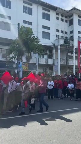 The video captures a lively street scene in Delkanda, Sri Lanka, during the daytime under bright, sunny weather. A large crowd of people is visible on the street, many of them dressed in red shirts and hats, indicating a coordinated gathering or protest. Several large red flags or banners are displayed along the street, possibly affiliated with the group.

The crowd is in motion, with people walking and mingling, suggesting a march or rally. In the foreground, a vendor is present, selling corn and other snacks from a cart, shielded by an orange umbrella. The street is lined with multi-story buildings, typical of an urban or semi-urban area. One building features a sign with "MADUZA" clearly visible. Other signs are too distant or blurry to read clearly, though one banner in the distance appears to contain Sinhalese script, possibly including "වෙලඳ" (Velanda, meaning trade or market).