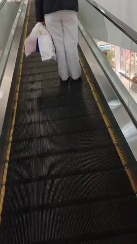 A person is riding an escalator upward in what appears to be a shopping mall. The person is wearing a dark jacket and light-colored pants, carrying multiple shopping bags. The escalator has metal sides and a black ribbed surface with yellow edges. Other stores and mall features are visible in the background. The camera angle is slightly above and behind the person, following them on the escalator.