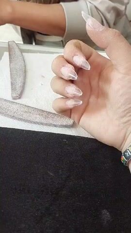 The video shows a person’s hand with long, clear acrylic nails, seemingly being prepped for further nail art. There are two nail files visible on a white surface, and the hand rests on a black surface. Another person, possibly a nail technician, is partially visible in the background, working on the nails. The nails have a slightly textured surface, possibly from filing. The overall scene suggests a nail salon or at-home manicure setting. The person’s hand has a bracelet with a colorful design.