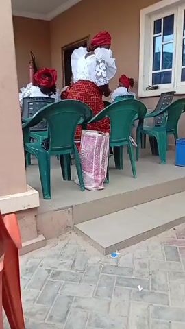 The video shows a gathering of people outdoors. Several women wearing traditional attire, including white headwear and patterned skirts, are sitting on green plastic chairs on a porch. A drum is visible next to one of the chairs. The camera pans to show a larger group of people seated under a green awning in what appears to be a courtyard. They are seated on blue, green, brown, and orange plastic chairs. The ground is paved with gray bricks. The setting appears to be a social gathering, possibly a meeting or celebration.