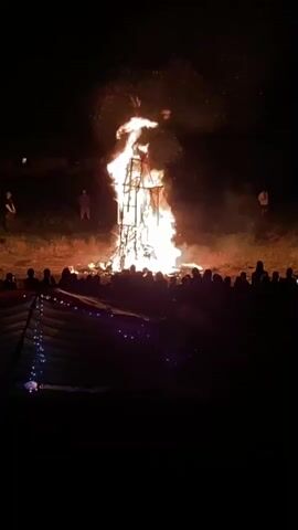 A large bonfire in the shape of a human effigy burns brightly at night. Sparks and flames erupt from the top and sides of the structure. A crowd of people, silhouetted by the fire, watches from a distance. In the foreground, a tent with string lights is visible. The ground around the bonfire appears to be covered in dry grass and dirt. The sky is dark, indicating it is nighttime.