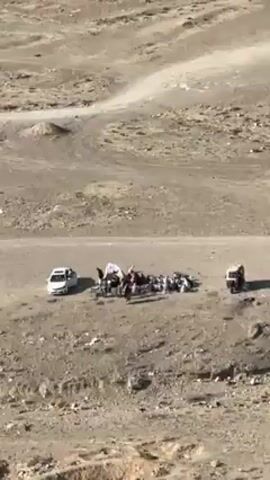 The video opens with a wide shot of a barren, mountainous landscape. In the foreground, a group of people are gathered near a white car and a small, three-wheeled vehicle. They appear to be interacting with an assortment of items spread on the ground. The camera then tilts upwards to reveal a broader view of the area, showing a city in the distance under a clear blue sky. The foreground is dominated by rocky terrain, providing a high vantage point over the surrounding landscape.
