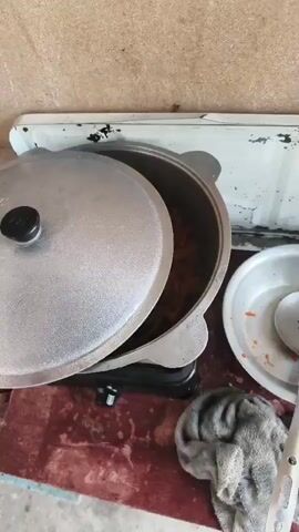 The video shows a close-up, overhead shot of a large, silver pot with a lid partially ajar, sitting on a small black stove. Inside the pot, chopped carrots and other ingredients are being fried in bubbling oil. The pot is placed on a rustic, reddish-brown surface that appears to be a table or countertop, which is set against a beige, textured wall. To the right of the pot, a white metal surface, possibly part of a larger stove or counter, is visible. A white bowl containing some of the cooked ingredients and a slotted spoon is also present on this surface. A grey, textured cloth is draped over the edge of the reddish-brown surface in the foreground. The scene suggests a cooking process in an outdoor or utilitarian setting.