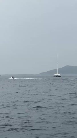 A jet ski moves across choppy, grey water, leaving a white wake. A large white sailboat with a single mast and dark hull is further out on the water, moving in the same direction. In the background, a hazy, green, mountainous island with buildings visible along its coast can be seen. The sky is overcast and grey.