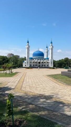 A mosque with a blue dome and two tall white minarets stands under a clear blue sky. A paved walkway leads to the entrance of the mosque. A few people can be seen walking in front of the mosque. Trees and some buildings are visible in the background. The sun is shining, casting shadows on the walkway.