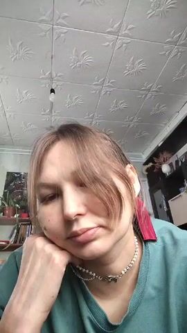 A young woman is sitting with her chin resting on her hand. She has light brown hair with bangs, a fair complexion, and is wearing a light green shirt and a pearl choker. She is looking directly at the camera with a neutral expression. The background shows a room with a white textured ceiling, a bookshelf, and a piece of artwork.