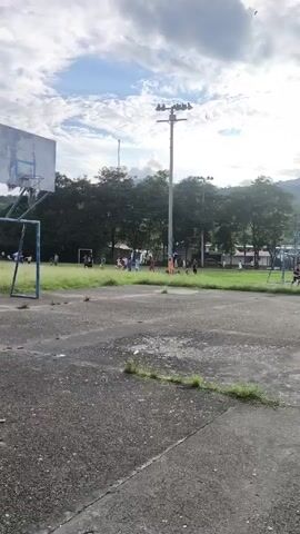The video shows a soccer game being played on a grassy field, viewed from behind a concrete basketball court. There are approximately 20 people, mostly dressed in athletic wear, spread across the field. Some are actively playing soccer, while others are standing on the sidelines. The field is bordered by trees and buildings. Behind the trees are mountains. A tall light pole stands between the basketball court and the soccer field. The sky is partly cloudy.