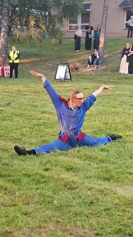 A person in a blue jumpsuit with red fringe on the shoulders performs a split on a grassy area. The person is wearing sunglasses and has their arms raised. In the background, there are several people watching the performance, some standing and some sitting on the grass. There is a building and trees in the background. There are also some objects in the background, including a sign and a red box. The lighting suggests it is daytime, possibly late afternoon due to the long shadows.