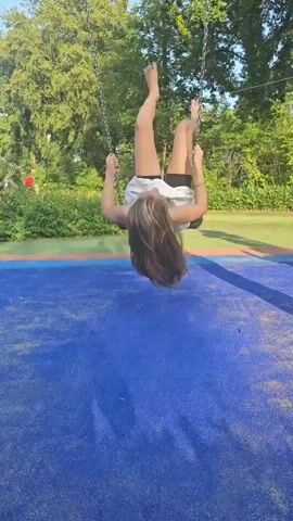 A young girl with long, wavy blonde hair is sitting on a swing at a playground. She is wearing a green tank top and a white eyelet skirt over black shorts. The swing set has metal chains and a black rubber seat. The ground beneath the swing is a blue rubber mat. In the background, there are green trees and bushes, suggesting an outdoor setting during the daytime, likely in warm weather. The girl appears to be enjoying herself, as she is smiling and touching her hair. The overall mood is playful and carefree. The location is identified as Groenlo, Netherlands.