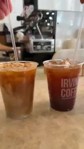 The video shows two hands stirring iced coffee beverages in clear plastic cups on a countertop. On the left is a lighter-colored drink, likely an iced latte, with a distinct layered appearance. On the right is a darker, brown coffee with the text "IRVIN COFF" printed on the side. In the background, an espresso machine is visible, along with a portion of a menu board or pricing list hanging on the wall. The countertop has a speckled beige pattern. Both hands are holding clear plastic straws and are in the process of stirring the beverages.