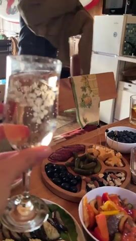 The video captures a close-up view of a hand holding a champagne flute filled with a bubbly, clear liquid. In the background, several other hands are visible, also holding drinks, suggesting a toast or celebration. A green glass bottle, likely beer, is prominently featured next to the champagne flute. A wooden platter laden with various appetizers, including olives, pickles, cheese, and cured meats, sits on a table in the foreground. The environment appears to be a casual indoor setting with hints of decor like a surfboard and artwork on the wall. The focus is on the drinks and food, implying a social gathering.