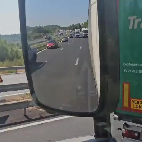 A truck's side mirror reflects a highway with traffic. A gray pickup truck with a black roll-up tonneau cover is seen driving in the right lane. The pickup truck has a license plate that reads "2-JFS-191". The highway is surrounded by green vegetation and hills under a clear sky. Another side mirror reflects more traffic and the road ahead.