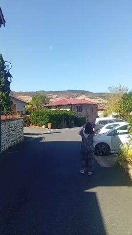 The video shows a person in a patterned jumpsuit walking away from the camera down a paved road. The person is walking towards a stone house with a red tile roof, which is partially obscured by dense green bushes. Behind the house, rolling hills covered in trees and fields are visible under a clear blue sky. Two cars are parked on the right side of the road. A trash can is near the house.