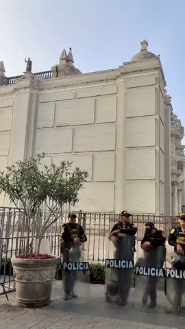 The video shows a line of police officers standing behind transparent shields that read "POLICIA" in yellow lettering. They are dressed in dark uniforms and caps, and several hold helmets. They are standing in front of a black iron fence with a large, white stone building in the background. A small tree in a stone pot is to the left of the officers. The sky is a clear blue.