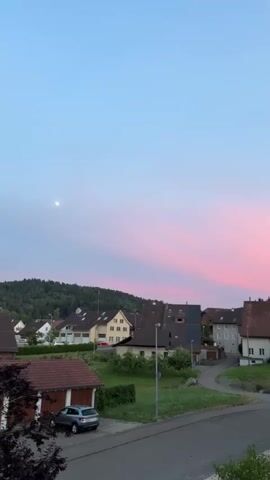 This is a vertical video of a picturesque village at dusk. The sky is a gradient of soft blues and pinks, with wispy clouds streaking across, suggesting a calm and beautiful sunset. A sliver of moon is visible in the upper left quadrant of the sky.

The scene depicts a European-style village with several houses, many featuring traditional tiled roofs. The architecture suggests a rural or suburban setting. Greenery, including trees and manicured lawns, is interspersed among the buildings. A road winds through the village, and a silver car is parked in front of a small building with a red-tiled roof in the foreground. Streetlights are visible, hinting at the transition from day to night. The overall impression is one of peacefulness and tranquility.

The time of day is clearly evening, as indicated by the sunset colors and the presence of the moon. The weather appears clear and pleasant. The location is identified as Lufingen, Switzerland, which aligns with the architectural style and the verdant landscape. There are no people visible in the frame, and no text is present. The camera is positioned at an elevated viewpoint, looking down onto the village. The mood conveyed is serene and idyllic.