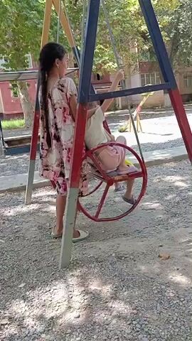 A girl with long dark hair in a braid, wearing a white and pink floral dress, stands to the left of a circular swing. She is looking at a second child who is seated in the swing, facing away from the camera. The child in the swing is wearing pink shorts and a light-colored shirt. The swing is red and appears to be made of metal. The background shows a playground with a gravel surface and trees. The swing set is made of metal poles painted red, blue, and yellow.