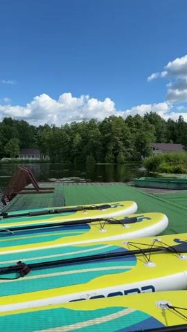 The video pans across a dock on a lake, showcasing several yellow and blue paddleboards lined up and ready for use. The lake is calm and reflects the blue sky with scattered white clouds. Dense green trees border the far side of the lake, with a few houses nestled among them. The dock is covered with a green mat.