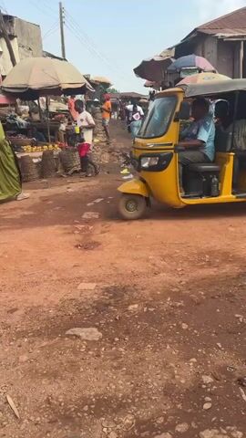 A yellow three-wheeled auto-rickshaw is moving on a dirt road. The auto-rickshaw has two passengers in the back and a driver in the front. The driver is wearing a blue and white shirt. The auto-rickshaw is passing by a market with various stalls and vendors. There are people walking around and shopping. The market appears to be in an outdoor setting with some stalls covered by umbrellas. The ground is dusty and reddish-brown. In the foreground, a portion of a silver pickup truck is visible. The overall scene depicts a busy market environment in what appears to be a developing country.