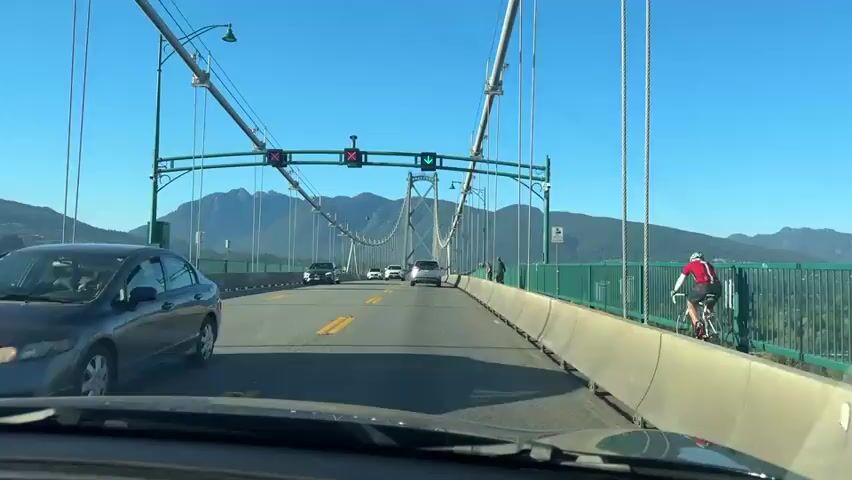 A first-person perspective video shows a car driving across a suspension bridge on a clear, sunny day. The bridge has multiple lanes of traffic with yellow dividing lines. Several other vehicles, including cars and what appears to be a motorcycle with a rider, are visible on the bridge. In the distance, mountains covered in trees can be seen under a bright blue sky. The bridge itself is characterized by its tall suspension towers and cables, as well as green railings along the sides. There are also traffic lights hanging over the lanes. The camera appears to be mounted on the dashboard or windshield of the car.