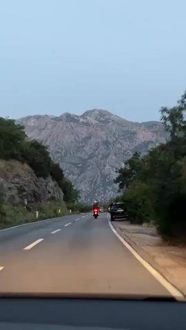 The video shows a road winding through a mountainous landscape. The sky is overcast, and the time appears to be dusk. The road has two lanes, with a white line dividing them. On the left side of the road, there is a steep, rocky slope covered in vegetation. On the right side, there is a parking area or shoulder where several vehicles are parked, including a black SUV and a white car. A motorcycle with a red taillight is traveling on the road ahead, in the same direction as the camera. The camera is positioned inside a vehicle, likely looking out the windshield. The overall scene is serene, with a sense of travel and exploration.