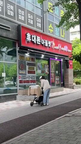 A woman wearing a white sweater, light blue jeans, a white baseball cap, and a face mask stands on a sidewalk, attending to a black baby stroller. A cream-colored and white jacket is draped over the top of the stroller. The stroller has four wheels. The background features a building with a red sign above it and advertising posters visible in the windows. The sidewalk is gray and brick-like.  The woman is facing the stroller and adjusting the jacket.