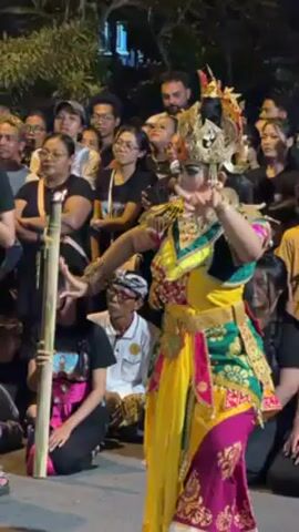 A woman is performing a traditional Balinese dance for a crowd of onlookers. The dancer is wearing an elaborate costume with vibrant colors, including yellow, green, and pink, and a golden headdress. She has long black hair and is using intricate hand gestures as part of her performance. The crowd is gathered closely around her, watching intently. Some people in the crowd are holding up their phones to record the dance. In the front of the crowd, a person is holding a bamboo instrument. The performance takes place outdoors at night, with artificial lighting illuminating the scene.