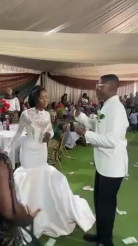 The video shows a wedding reception with the bride and groom dancing on a green carpet. The bride wears a white, fitted dress, and the groom is in a white suit jacket and black pants. The groom throws money in the air, and it scatters around them on the carpet. Guests are seated in the background, watching the couple. The reception is held in a tent-like structure with a white ceiling and decorated with brown and white fabric draping.