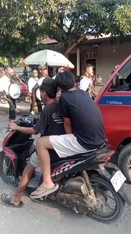 A group of people are gathered on a street, some playing musical instruments, likely a marching band. In the foreground, two young men are sitting on a motorcycle, one driving and the other as a passenger. The driver is wearing a black t-shirt and white shorts, with flip-flops on his feet. The passenger is wearing a black t-shirt and white shorts. The motorcycle is parked on a dirt road, with a red vehicle to the right and more people and musical instruments in the background. Trees and buildings are visible in the background, indicating an outdoor setting. The sun appears to be shining, casting shadows.