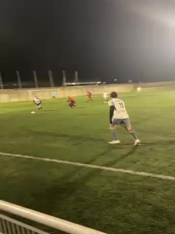 The video shows a soccer game being played at night on a green grass field. Players in red jerseys are playing against players in white and gray jerseys. The camera follows the action as the players move the ball down the field. There are floodlights illuminating the field. A fence is visible in the foreground of the video. Buildings can be seen in the background.