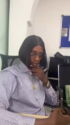 A person wearing a short black bob wig, glasses, a patterned light blue shirt, and a gold necklace is sitting in a black office chair. The person’s right hand is touching their chin, and they are looking slightly off camera. Behind them is an office desk with a computer monitor and what appears to be a newspaper in Chinese. A blue and white bulletin board hangs on the wall in the background. The setting appears to be a modern office space.