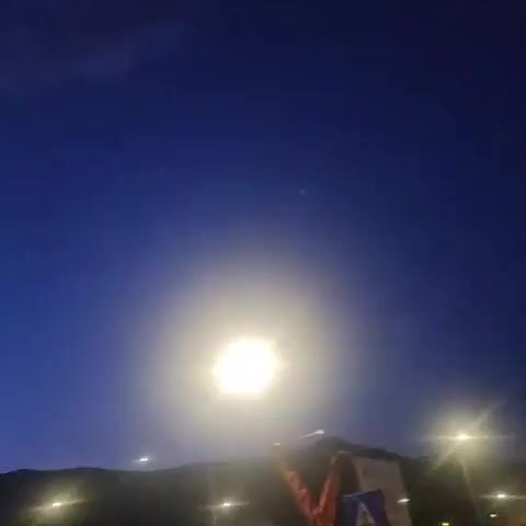 The video shows a dusky sky with a bright light, possibly a streetlamp or the moon, in the upper right quadrant of the frame. The sky transitions from deep blue at the top to a lighter purple hue near the horizon. Silhouetted against the sky are mountains and a line of lights, likely streetlights or building lights. In the foreground, parked cars are visible under a blue awning. The image is slightly blurry and appears to be taken from a moving vehicle.