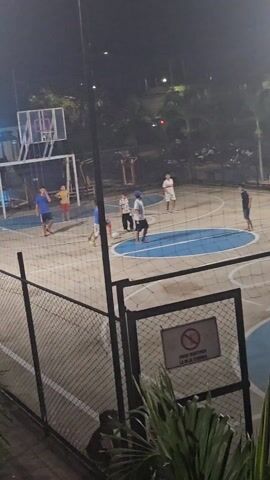 At night, a group of boys are on a basketball and soccer court surrounded by a chain-link fence. The court is painted with light blue circles and lines on a tan surface. A soccer goal is visible near a basketball hoop. A sign on the fence in Spanish reads "Favor mantener la reja cerrada," which translates to "Please keep the gate closed." Some of the boys are kicking a soccer ball around the center circle of the court. A few palm trees and other vegetation are visible around the court. Parked motorcycles are seen behind the fence.