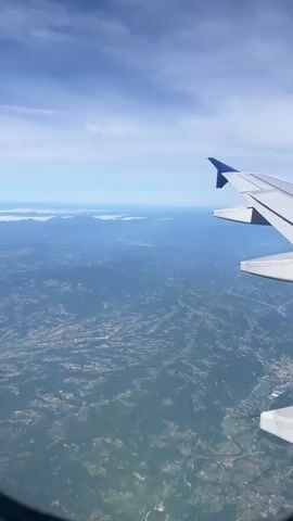 A clear, sunny daytime aerial view from an airplane window captures a vast landscape below. The white wing of the aircraft, with a blue accent on its wingtip, occupies the right side of the frame. Below, a diverse terrain unfolds, dominated by green and brown hues, indicative of hills, mountains, forests, and possibly agricultural fields. Small towns or settlements are visible as clusters of buildings scattered across the land. In the distant background, a lighter blue haze blends land and sky, with what appears to be a large body of water or a distant landmass extending towards the horizon. The sky above is bright blue with scattered white clouds, suggesting good visibility. The airplane is in flight, providing a dynamic perspective of the earth's surface. No visible text is present.