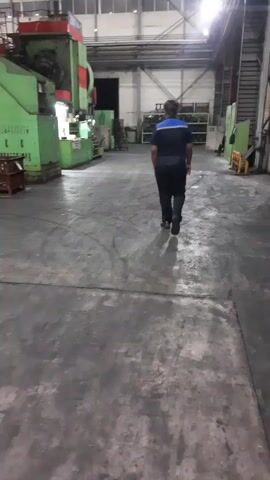 A person walks down a long, industrial hallway. The person is wearing a blue and black uniform with a blue stripe across the shoulders. The hallway is lined with large green and red machinery, some of which appear to be presses or stamping machines. Shelving units with various items are visible in the background. The floor is concrete and shows signs of wear, with tire marks and scuffing. Bright overhead lights illuminate the space, casting long shadows. The overall impression is of a manufacturing facility or workshop. The time of day appears to be daytime due to the lighting, but there are no windows visible to confirm. No weather conditions can be determined. The location is identified as Miryang-si, South Korea. There is no discernible text visible in the video.