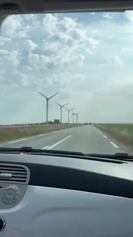 The video shows a view from inside a car driving on a road. The road is paved and has white lines marking the lanes. On the left side of the road, there is a field with several wind turbines lined up in the distance. The sky is cloudy with patches of blue. The dashboard of the car is visible in the foreground. The car is moving forward, and the scenery outside is passing by.