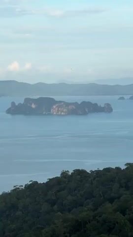 A static, long-shot video captures a serene tropical seascape under a bright, cloudy sky. In the foreground, a dense, dark green forest blankets a hillside, its lush canopy extending towards the bottom edge of the frame. Beyond the forest, a vast expanse of calm, blue water dominates the middle ground, dotted with several majestic limestone karst islands of varying sizes, their rocky formations rising dramatically from the sea. These islands are a deep, earthy brown, some topped with a scattering of green vegetation. In the far distance, a series of hazy, blue-green mountains recede into the horizon, creating a sense of depth and scale. The sky above is a pale blue, with scattered, fluffy white clouds adding texture and dimension. The overall atmosphere is one of tranquility and natural beauty, evoking a sense of remote, unspoiled paradise. There are no people or discernible objects in motion within the frame.