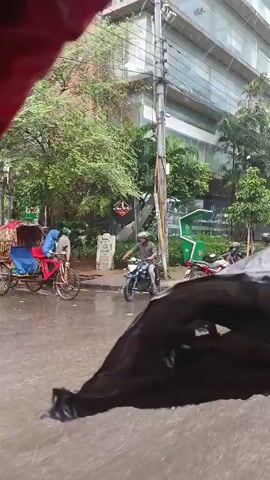 The camera captures a rainy day scene from what appears to be a moving vehicle. The view is partially obstructed by a black object, likely part of the vehicle's interior. The roadside features a row of parked motorcycles, lush greenery, and modern buildings. People are seen walking and standing near the buildings. A silver car passes by, followed by a black one. A cyclist with a red bicycle rides past in the opposite direction. The environment suggests an urban setting with a mix of commercial establishments and parking areas.