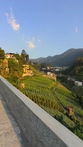 A slow pan across a mountainous landscape shows terraced farms with crops growing. Several buildings, including houses and what appear to be small sheds, are scattered throughout the scene. In the background, more houses and buildings are visible on the slopes of the mountains. The sky is blue with a few white clouds, and the sun appears to be setting, casting a warm light on the scene. The foreground shows a concrete barrier with a textured surface, suggesting the vantage point is from a road or walkway.