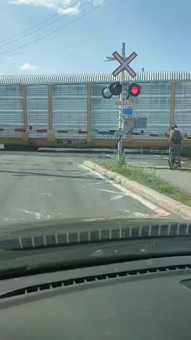 A first-person perspective video captures a train crossing a road. The camera is inside a vehicle, showing the dashboard in the foreground. A yellow train with metal siding and graffiti is passing by. A railroad crossing signal with flashing red lights is visible. A person on a bicycle is stopped on the right side of the road, waiting for the train to pass. The sky is blue with some clouds. The road appears to be asphalt, with a curb and some overgrown grass on the side.