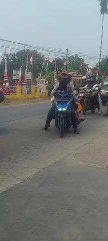 A group of people are riding on motorbikes, some are also in the back of a truck. There are Indonesian flags visible. One motorbike is blue, another is black, and another is dark colored. The people on the motorbikes are of various ages and genders. There is a large statue on top of the truck, resembling an elephant. The scene appears to be a parade or procession. The weather seems overcast.