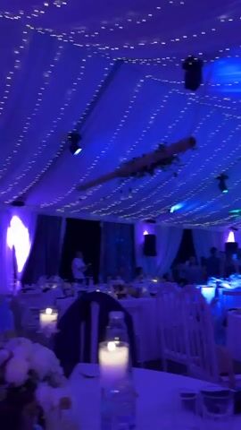 The video, shot from a low angle, captures the interior of a festive event tent, likely for a wedding or party. Several round tables, set with white tablecloths, glassware, and candles, are arranged across the space. White chairs with arched backs are placed around the tables. The ceiling of the tent is draped with white fabric and illuminated by numerous strings of fairy lights, creating a sparkling effect. Two large, dark cylindrical objects, possibly decorative elements or sound equipment, hang from the ceiling, along with what appears to be a rustic wooden beam adorned with some greenery. The lighting in the tent changes color, shifting from blue to green. People are visible in the background, some standing and mingling, others seated at tables, suggesting a lively atmosphere. At one point, a person can be seen near a stage or performance area, possibly a musician.