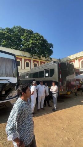 The video depicts an outdoor scene with several people standing and walking around. There are multiple buildings visible, with one featuring a white and red design, and another appearing older and made of grey stone. In the foreground, a group of men is standing near a large, dark bus-like vehicle. There are white tents set up in the area, with people gathered underneath them. The ground is a dirt surface. The sky is clear and blue.